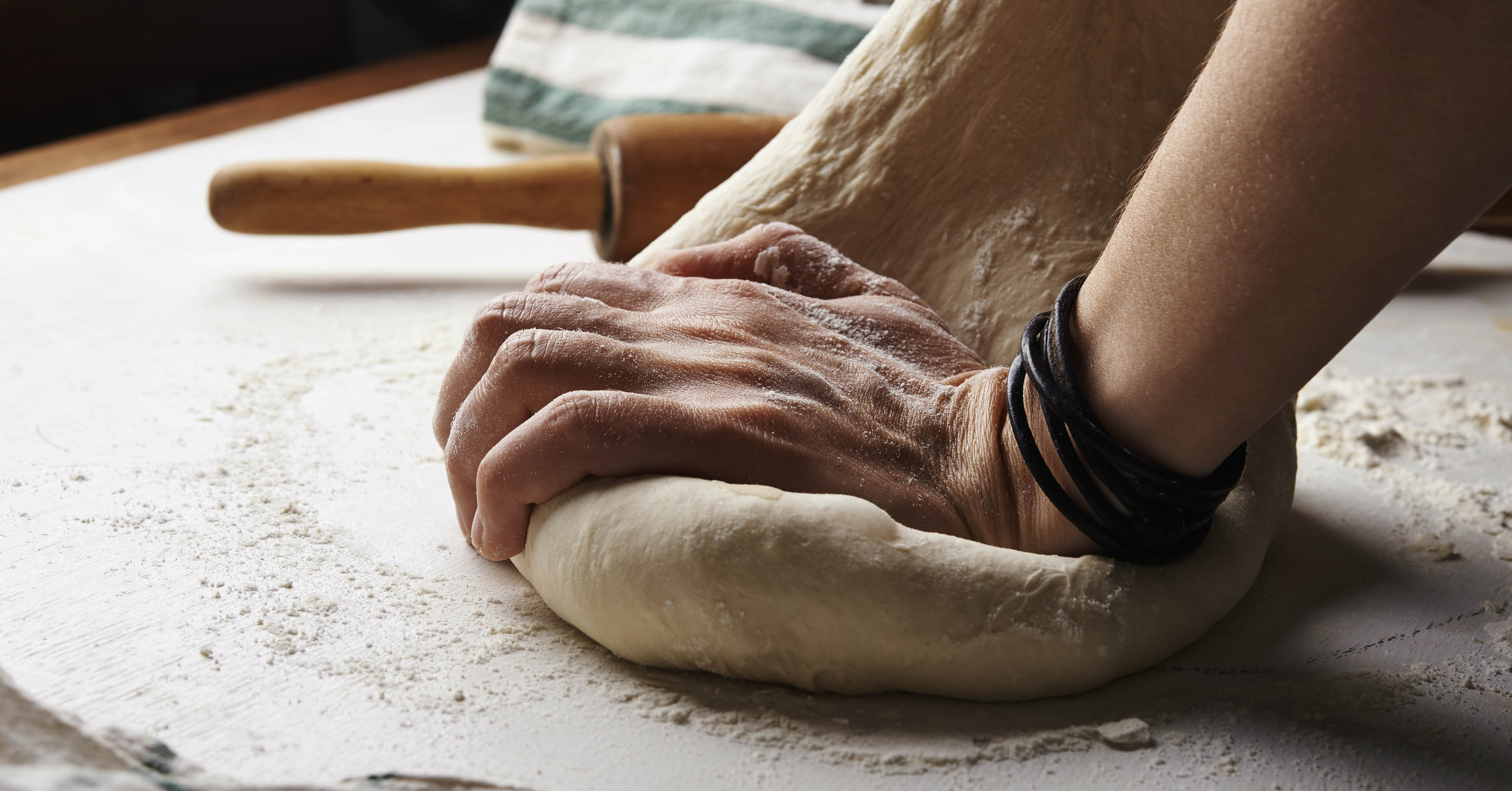 Close-up van een hand die deeg kneedt op een met bloem bestoven werkblad, met een deegroller en keukendoek op de achtergrond.