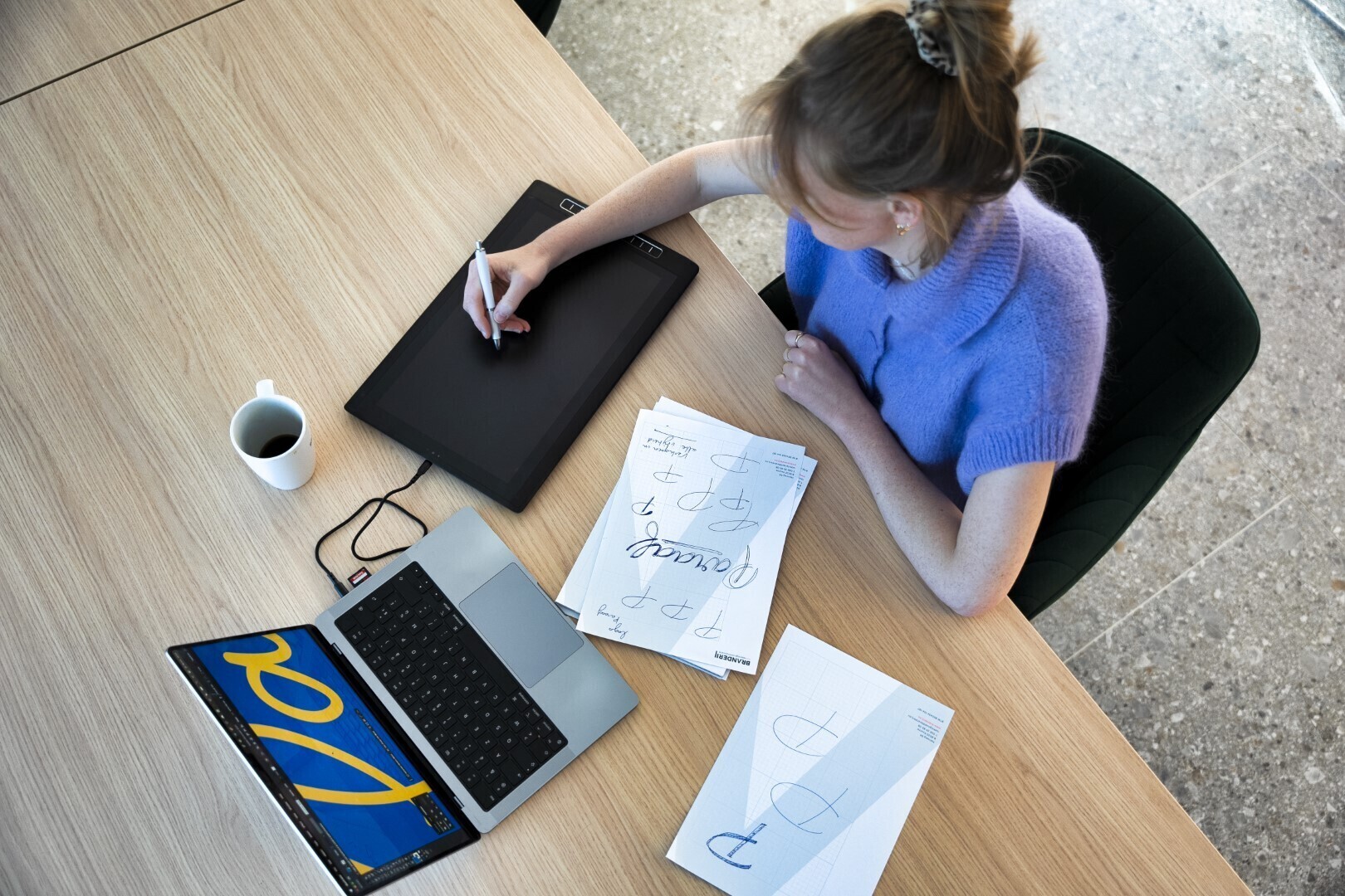 Designer werkt aan een logo-ontwerp met een tekentablet aan een houten bureau, omringd door schetsen, een laptop met het woord “Lora” op het scherm en een kop koffie.