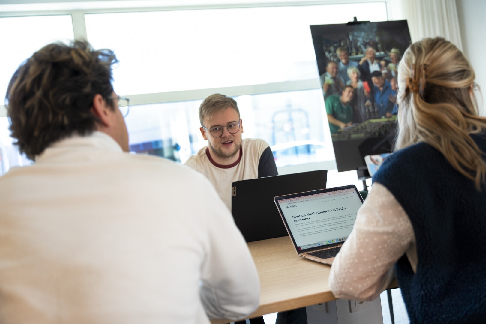 Drie collega’s in gesprek rond een tafel met laptops in een lichte kantoorruimte, met een kunstposter op een schildersezel op de achtergrond