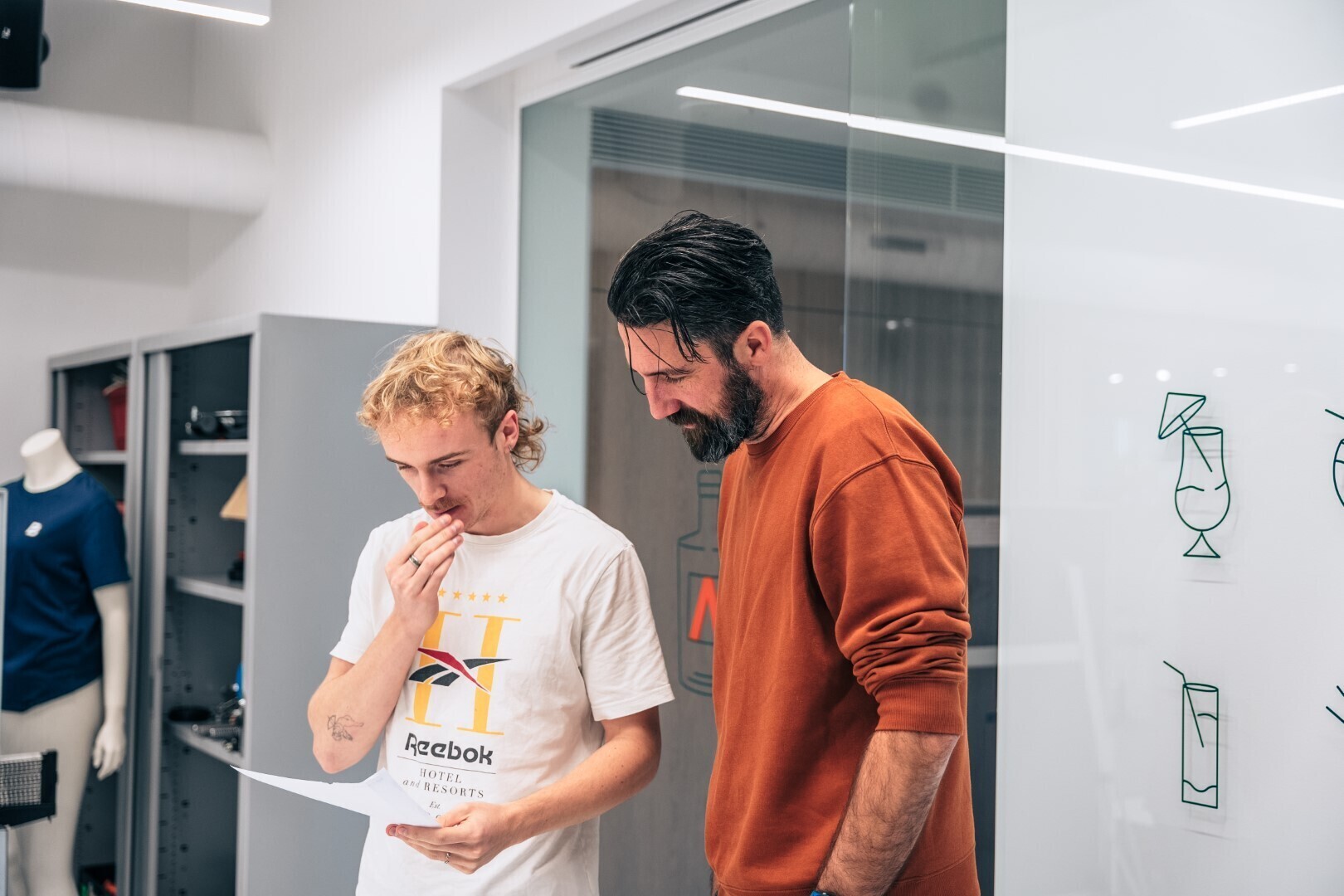 Twee mannen staan binnen voor een glazen wand en kijken geconcentreerd naar een document, in een moderne kantooromgeving.