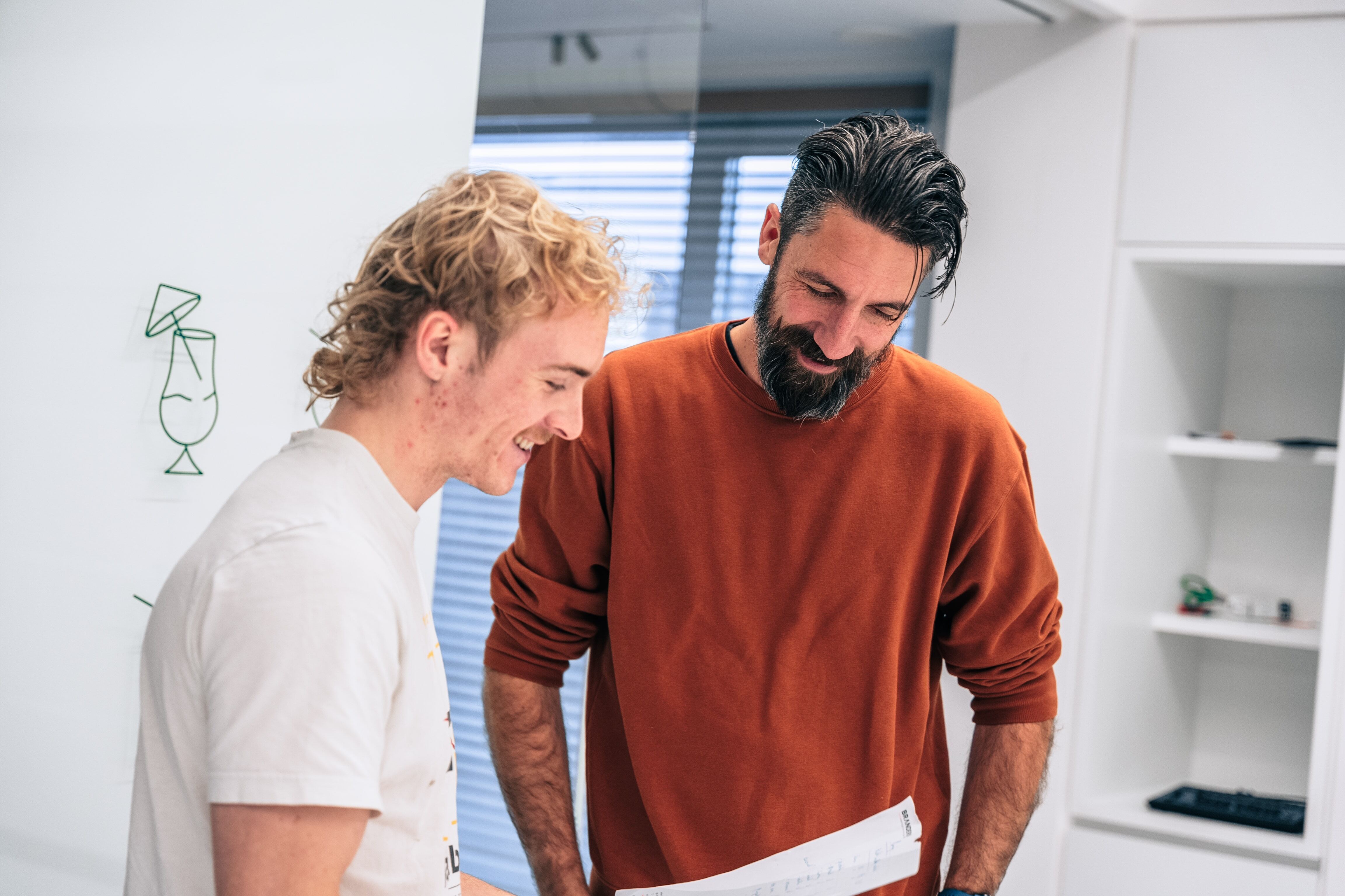 Twee collega’s staan naast elkaar te lachen terwijl ze samen een document bekijken in een moderne kantooromgeving met whiteboard en raam op de achtergrond