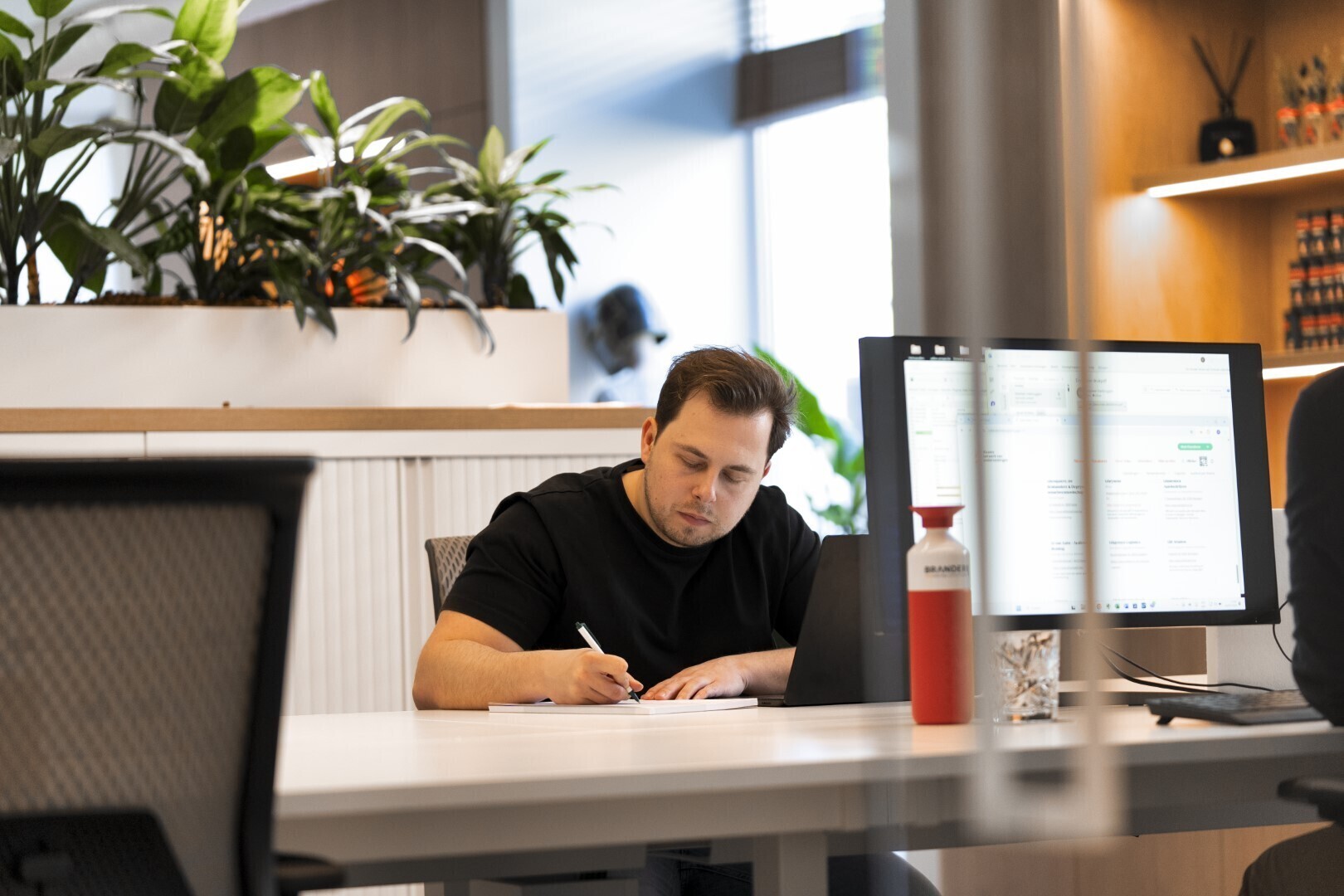 Man in zwart T-shirt werkt geconcentreerd aan zijn bureau in een moderne kantoorruimte met computerscherm, waterfles en planten op de achtergrond