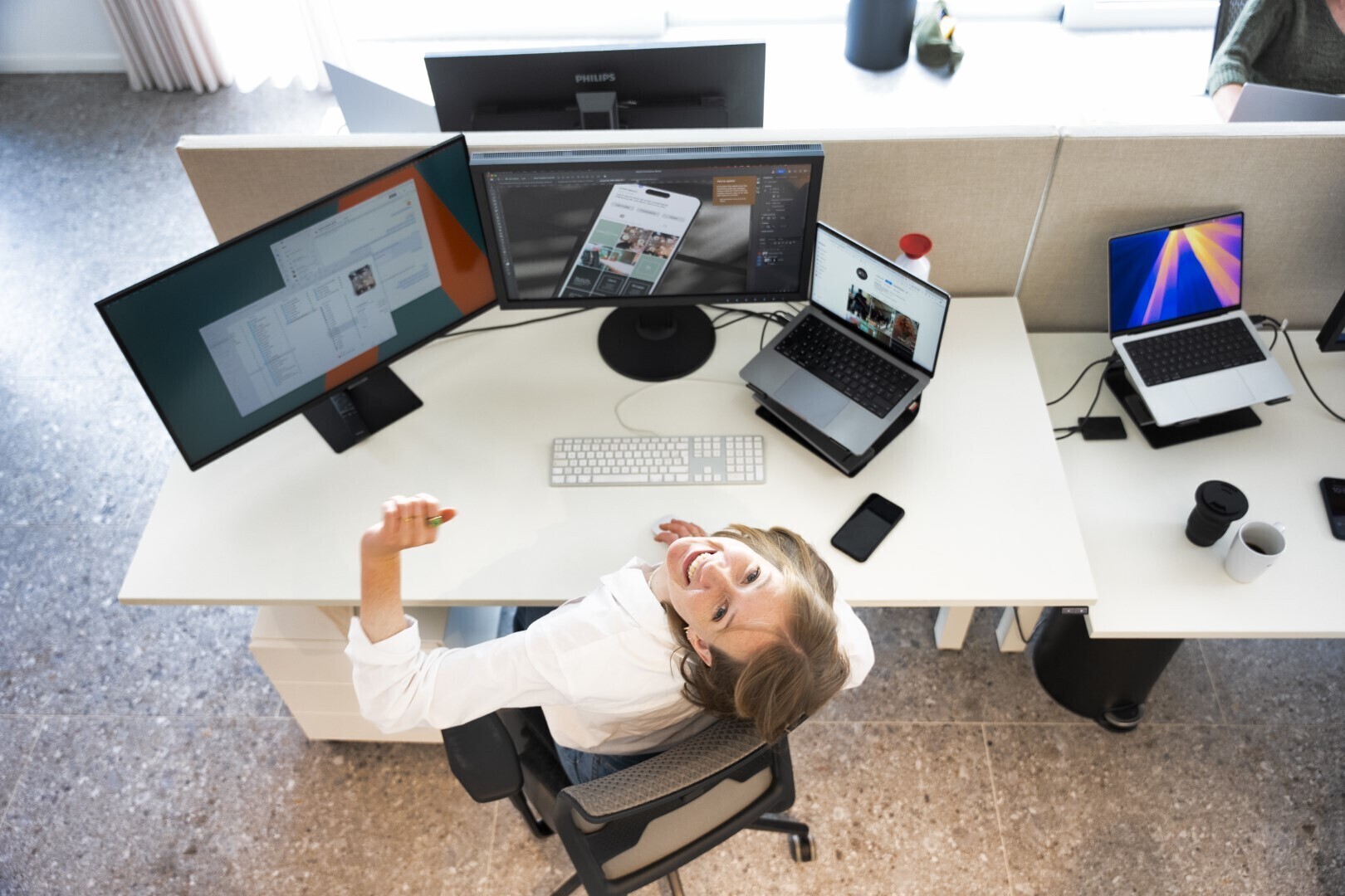 Vrouw aan een moderne werkplek met meerdere computerschermen, laptops en smartphones, kijkt glimlachend omhoog naar de camera vanuit haar bureaustoel.