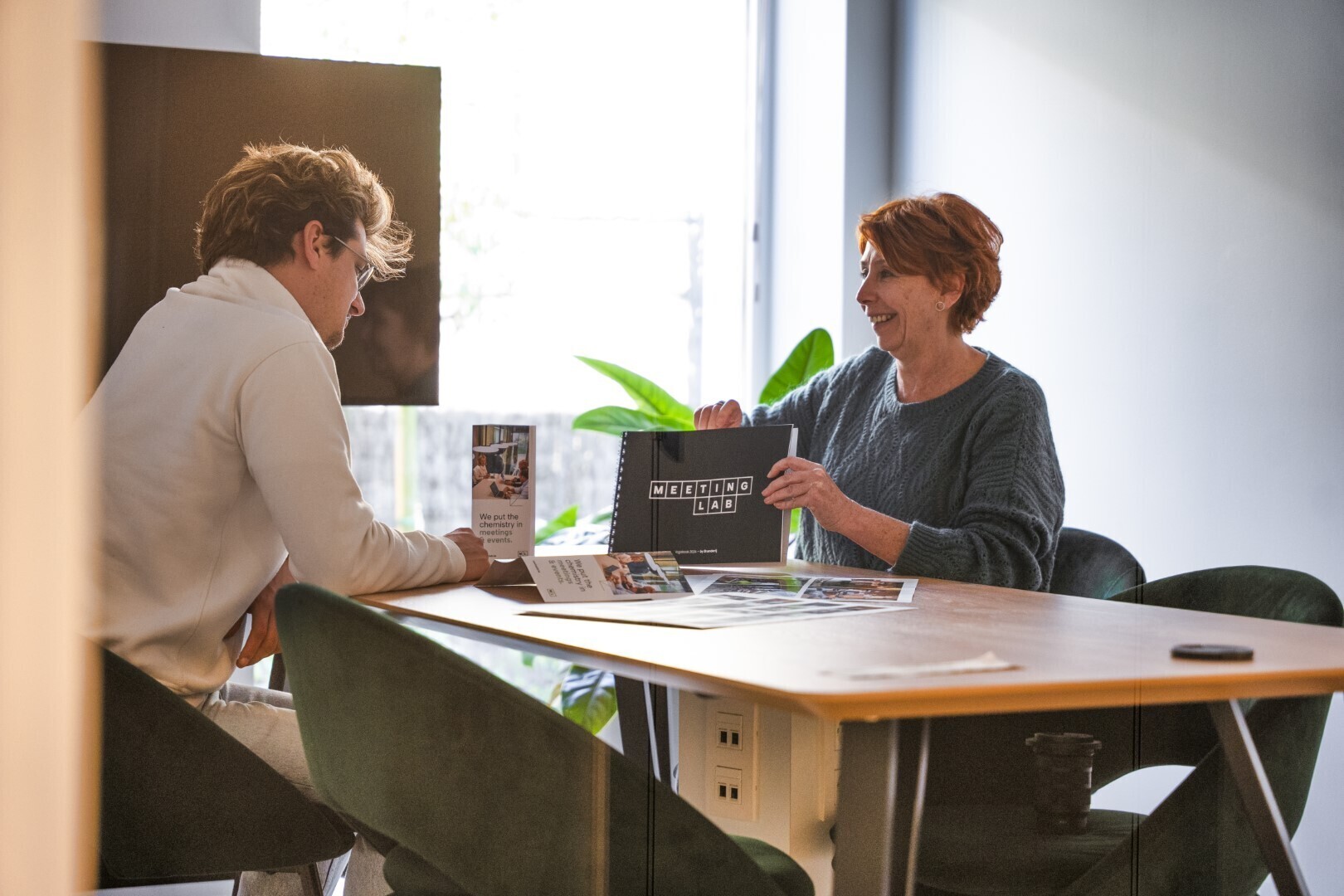 Man en vrouw zitten tegenover elkaar aan een vergadertafel in een lichte kantoorruimte en bespreken drukwerk en documenten tijdens een klantgesprek