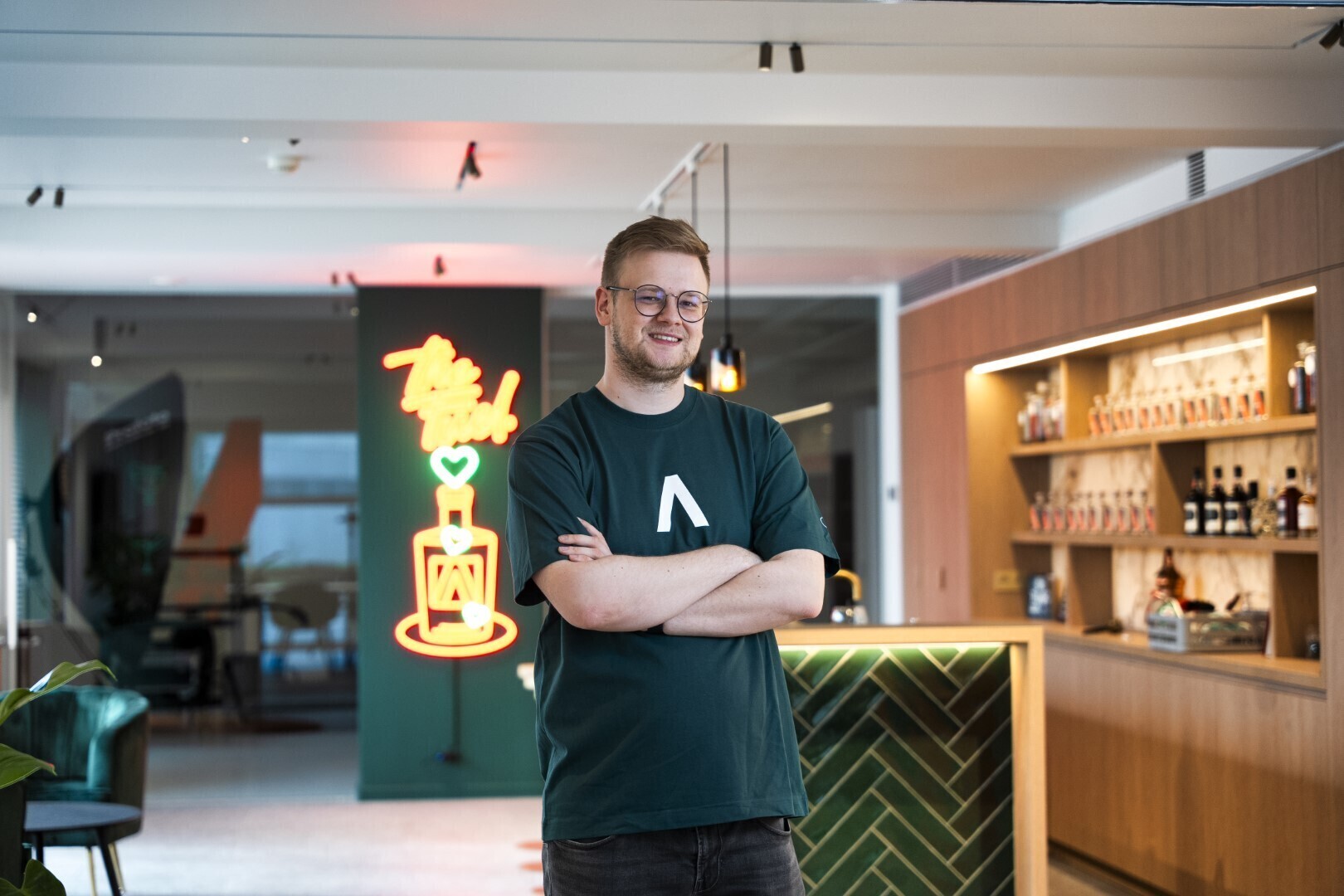Jonge man met bril en donker T-shirt met logo poseert met gekruiste armen voor een moderne bar met groene tegels en neonverlichting.