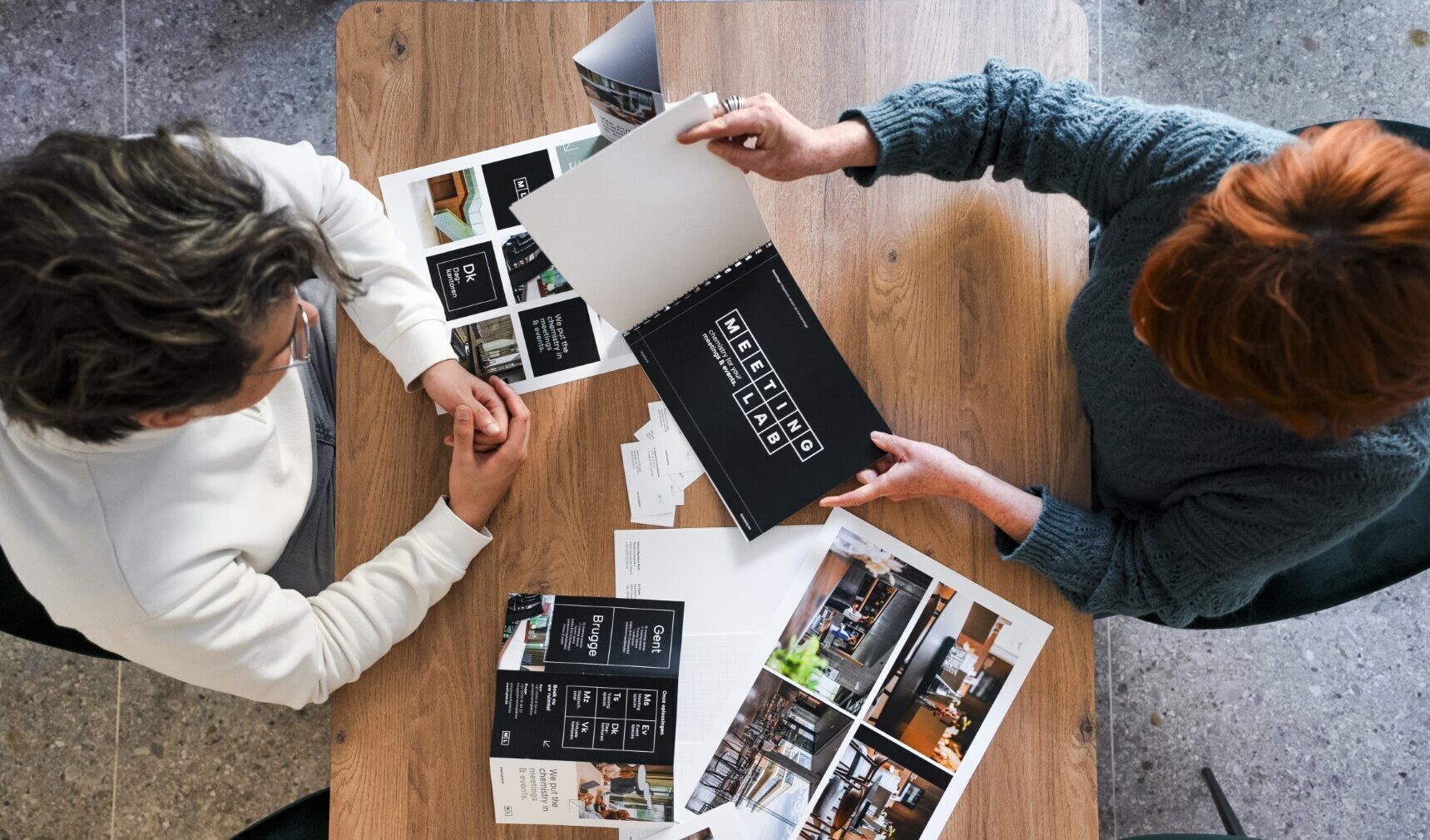 Bovenaanzicht van twee personen aan een houten tafel die samen een brandingpresentatie met drukwerk en moodboards bekijken.