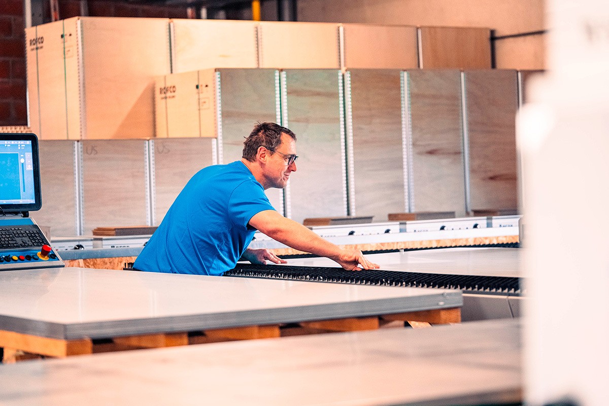 Man in blauw T-shirt werkt geconcentreerd aan een grote zaag- of freesmachine in een productieomgeving.