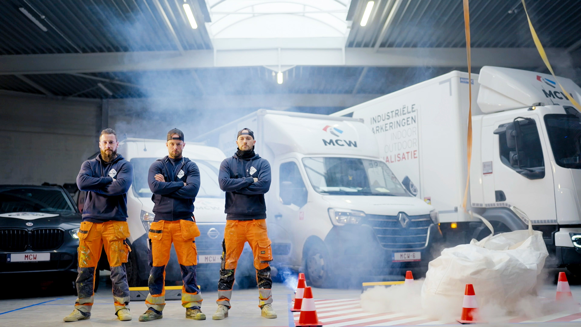 Drie mannen in werkkledij poseren stoer voor vrachtwagens van MCW in een loods, omringd door rook en kegels.
