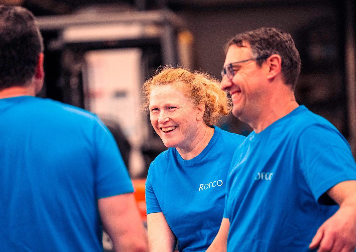 Drie collega’s in blauwe T-shirts met bedrijfslogo lachen samen in een werkplaats of productieomgeving.